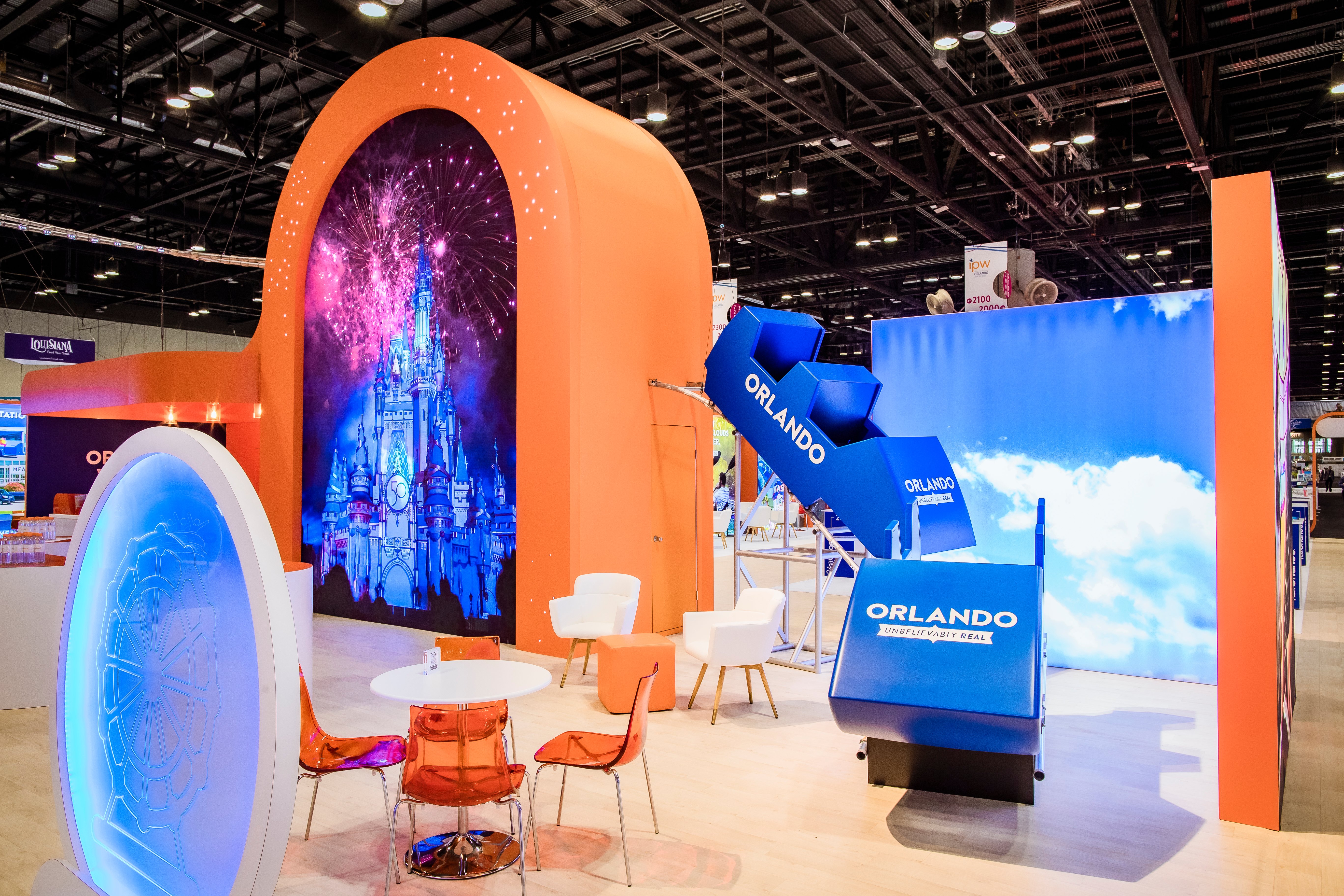 An image of the Visit orlando booth at IPW.  We can see a prop roller coaster, a table with orange chairs, in front of it,  and an orange arched wall with a moving image of the Cinderella Tower at Walt Disney World to the left. 