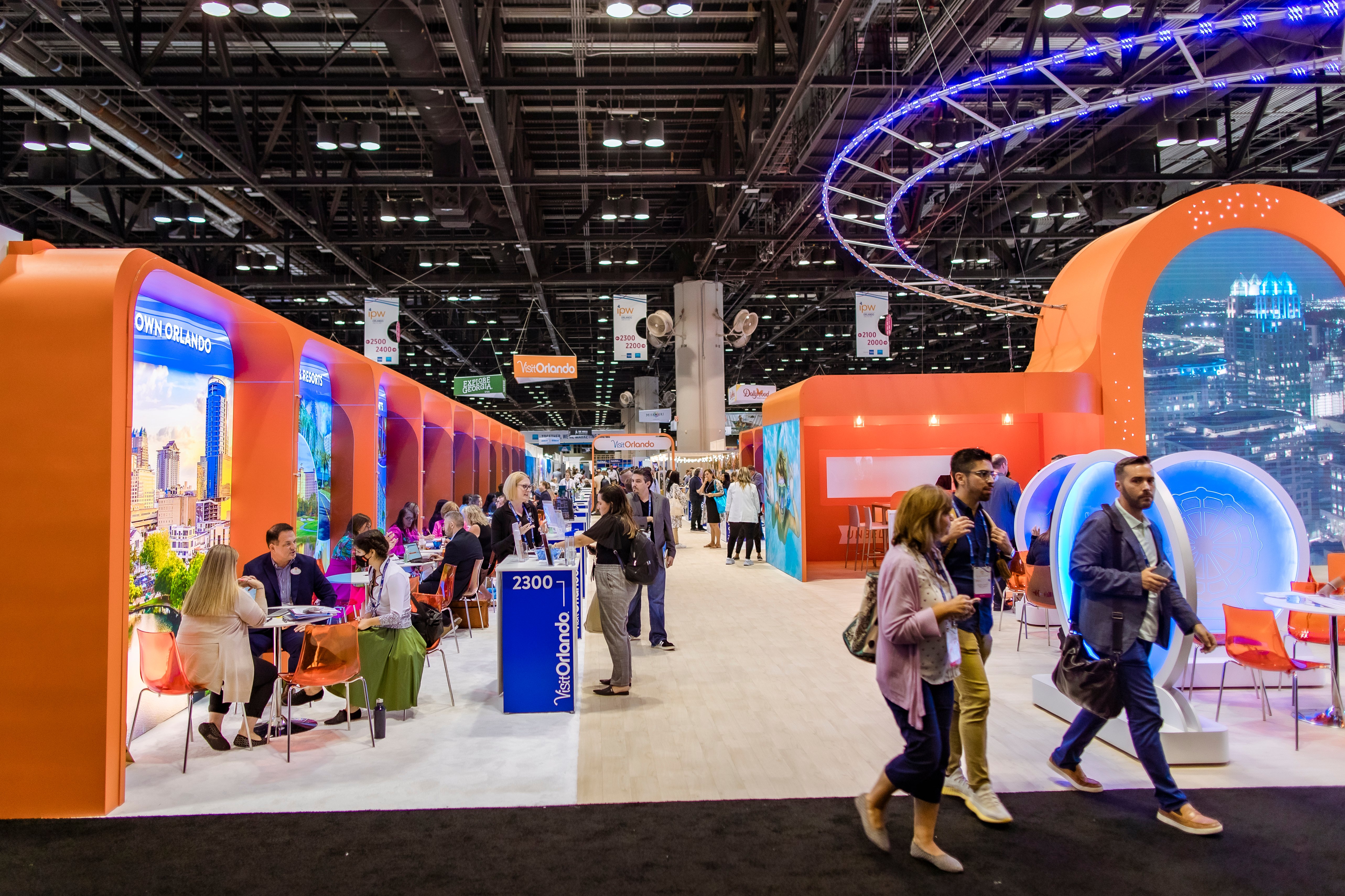 People walking around the visit Orlando booth.  We can see sets of tables and chairs,  each numbered. 