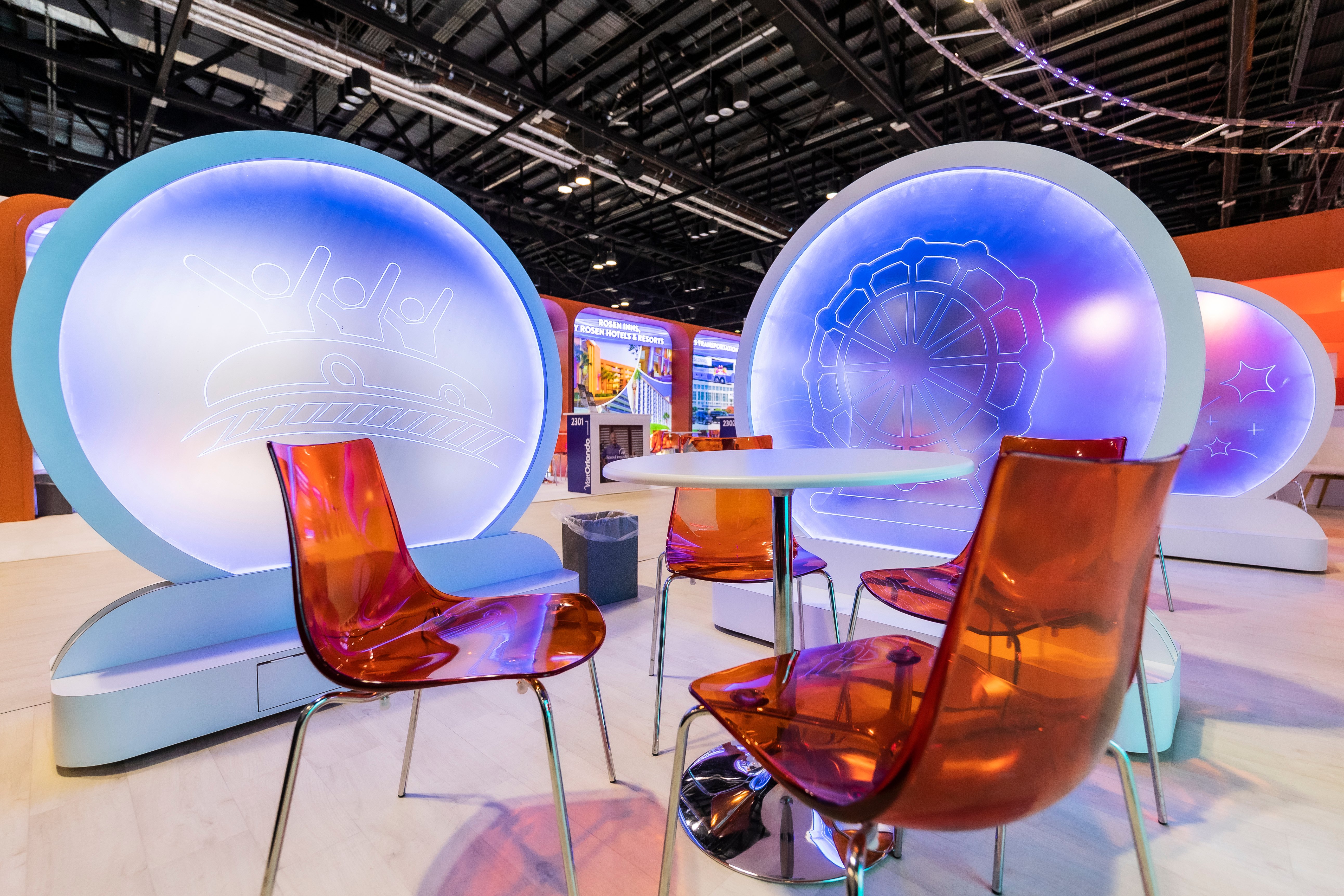 An image of the Visit orlando booth at IPW.  We can see a prop roller coaster, a table with orange chairs, in front of it,  and an orange arched wall with a moving image of the Cinderella Tower at Walt Disney World to the left. 