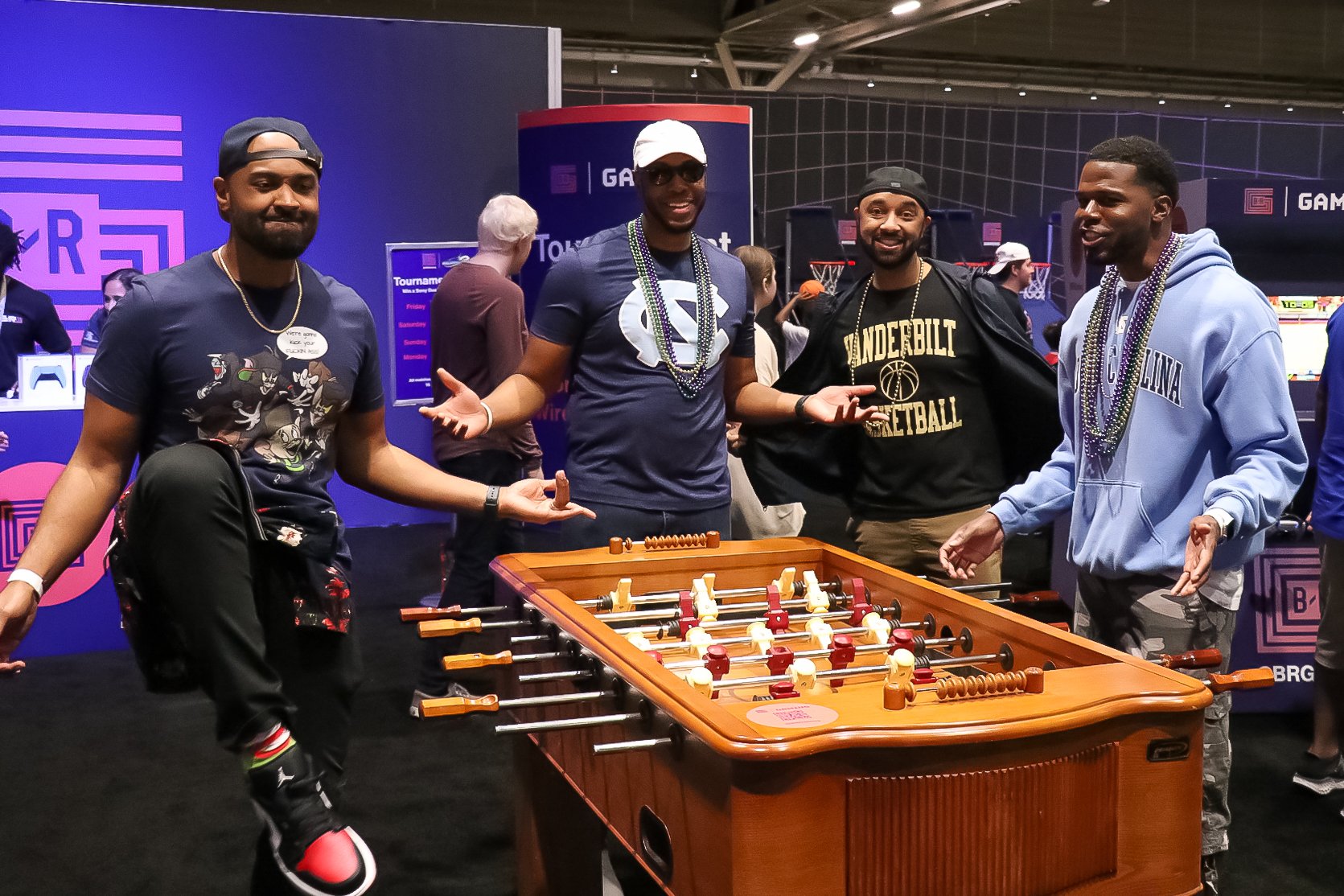 4 black men playing foosball pose for the camera. two wear North Carolina merch and one wears a Vanderbilt basketball shirt. 