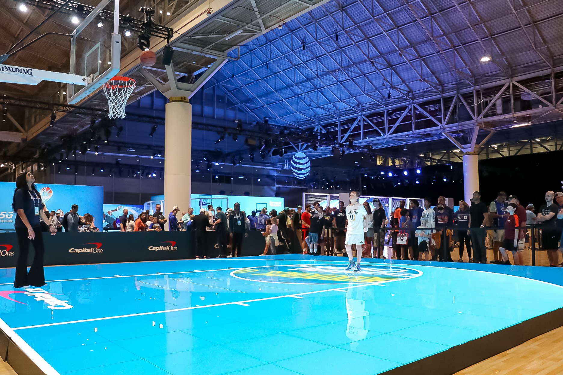 An image of a bright blue court sponsored by Capital One. A crowd of people is seen behind the court, along with other booths.