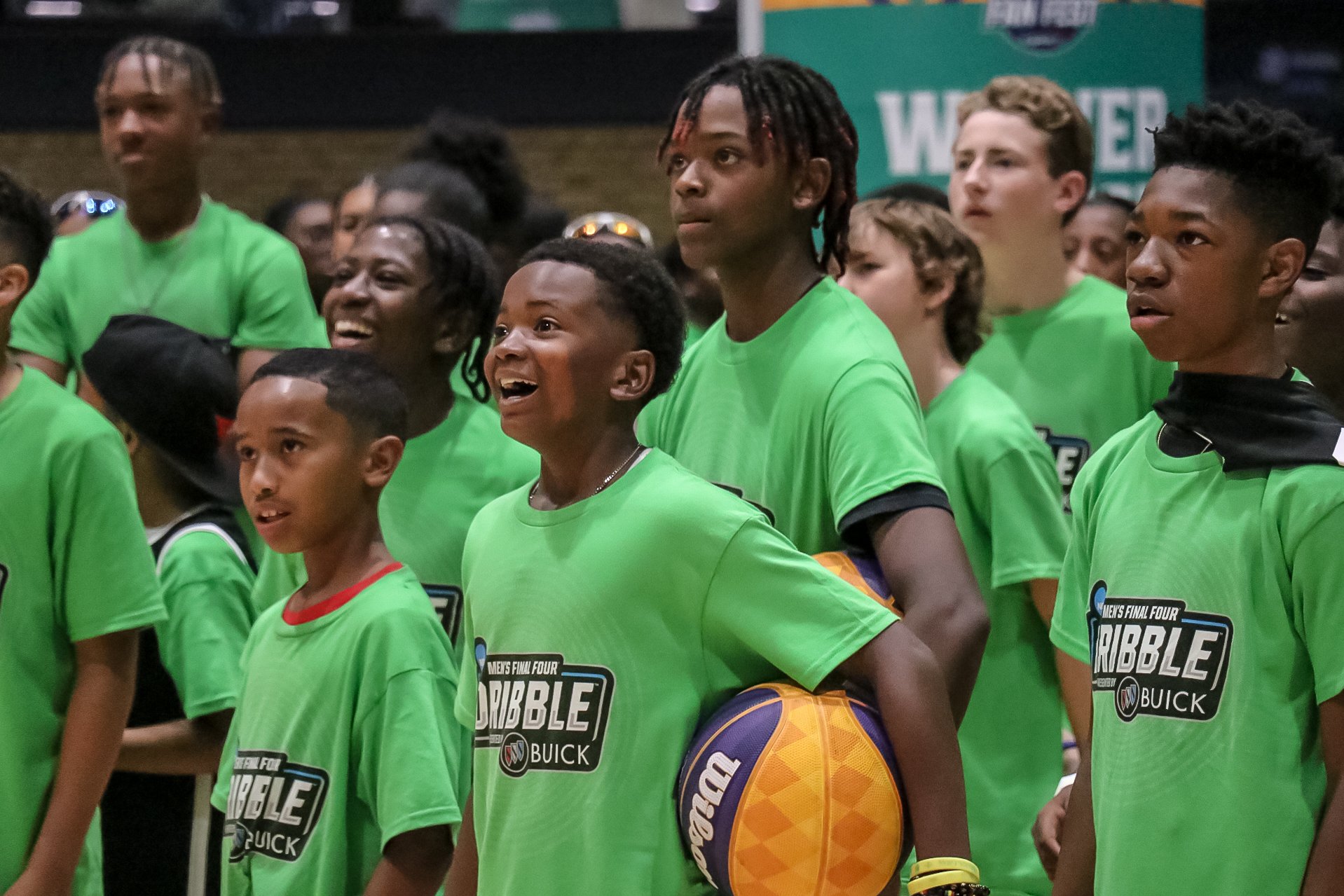 A group of young children  all wearing green shirts with a  dribble contest logo look to the left. Some smile and others look in amazement. 