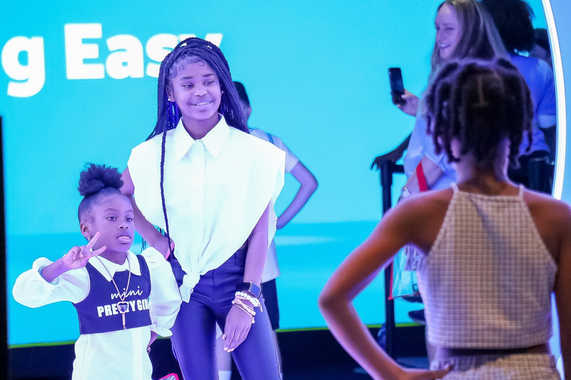 Two black girls, one around 2-4 and the other 8-11 pose together for a photo in front of a light blue LED display. 