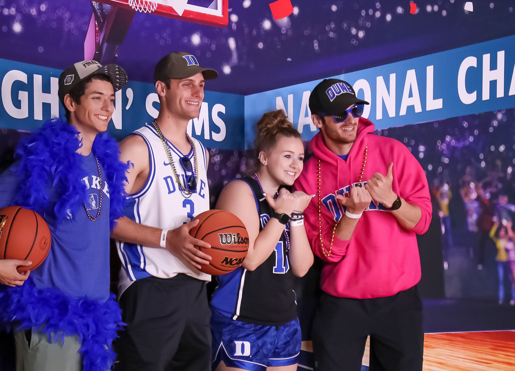 4 people wearing Duke swag pose for a photo.. The two on the left hold basketballs while the two on the right put their fists together in a sideways thumbs up. 