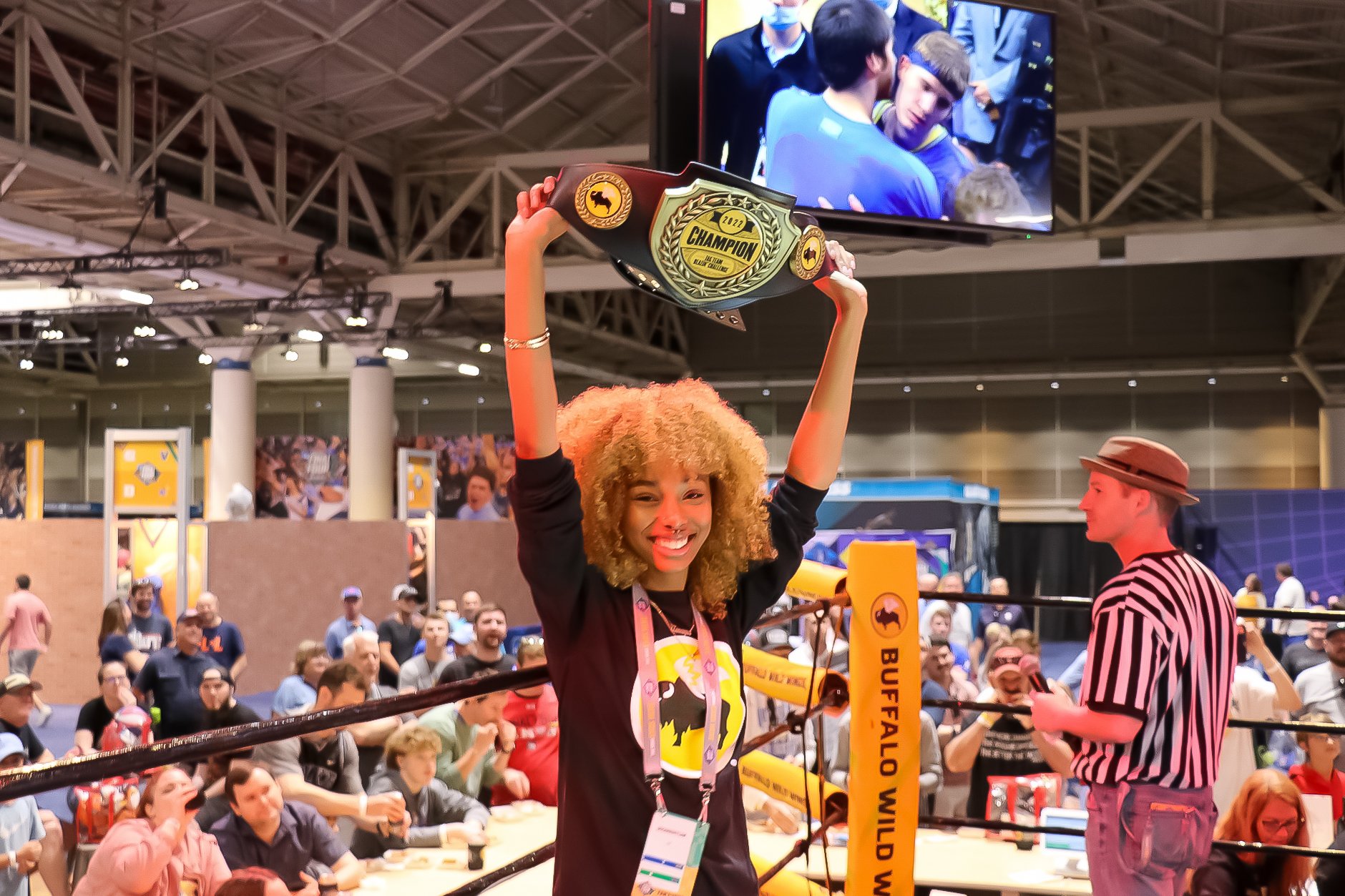 A black woman cheerfully holds up a wrestling belt at the Buffalo wild wings booth. Behind her is a small crowd. 